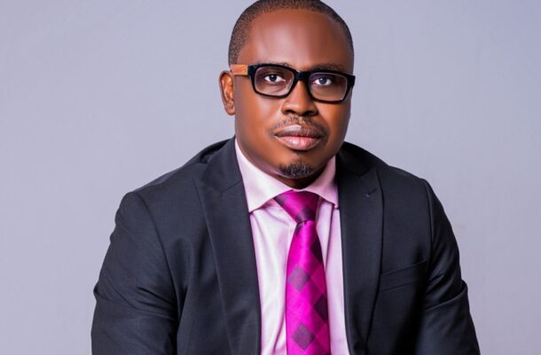 WhatsApp Image 2026-07-02 at 085009 | Ghanaguardian.com Portrait of a man in a dark suit with a pink shirt and bright magenta tie, wearing glasses against a gray backdrop.