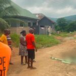Group of people in orange shirts gather on a dusty village street; a covered body lies blurred on the ground nearby.
