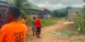 Group of people in orange shirts gather on a dusty village street; a covered body lies blurred on the ground nearby.