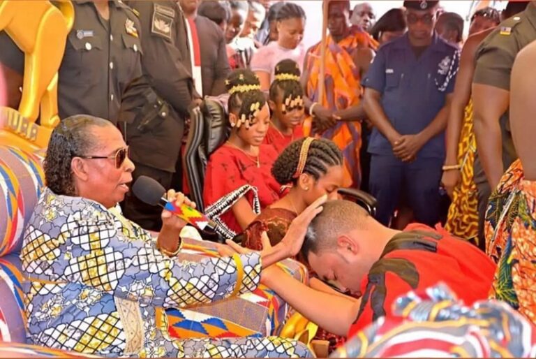 An elderly woman with a patterned dress speaks into a microphone while a man in a red shirt bows his head in front of her; a crowd watches in colorful attire.