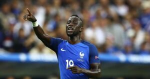 French soccer player in blue jersey number 10 points upward in celebration during a match, with a cheering crowd in the background.