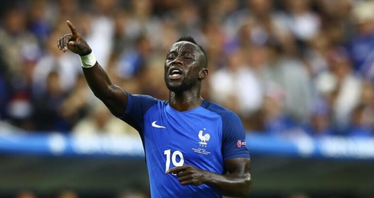 French soccer player in blue jersey number 10 points upward in celebration during a match, with a cheering crowd in the background.