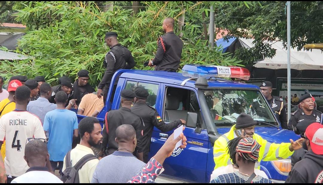 WhatsApp-Image-2026-07-11-at-043536-1 | Ghanaguardian.com Crowd gathers around a blue police pickup with officers in black uniforms and red-and-black shoulder cords near tents and trees.