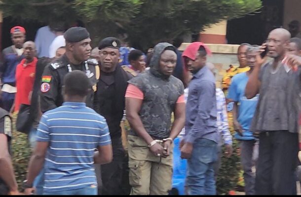 WhatsApp-Image-2026-07-11-at-043536-2 | Ghanaguardian.com Man in handcuffs escorted by uniformed officers as a crowd watches at an outdoor event.”,