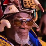 Older man with a gray beard wearing a colorful, ornate headdress and sunglasses.