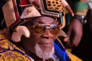 Older man with a gray beard wearing a colorful, ornate headdress and sunglasses.