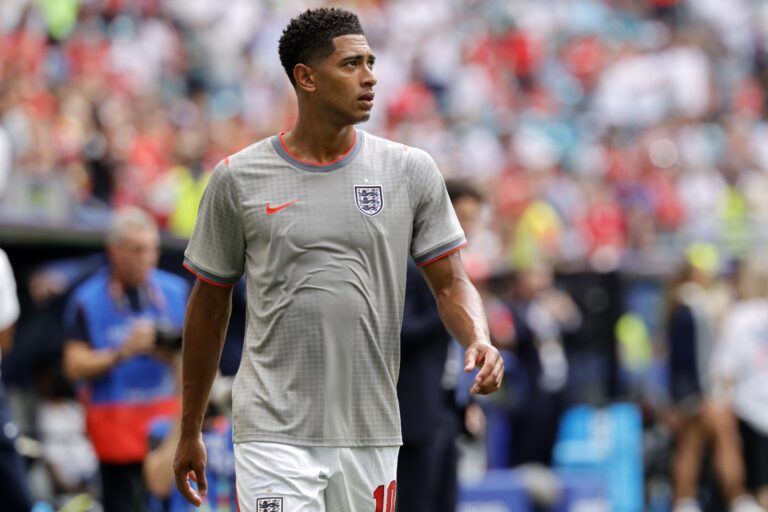 Footballer in a gray England jersey walking on the pitch with a blurred stadium crowd in the background, during a match.