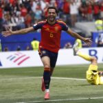 Spanish player in red jersey #6 celebrates with arms outstretched on the pitch as the goalkeeper lies on the ground behind him.