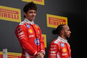 Two Ferrari drivers in red racing suits stand on a podium with yellow Pirelli signs behind them.