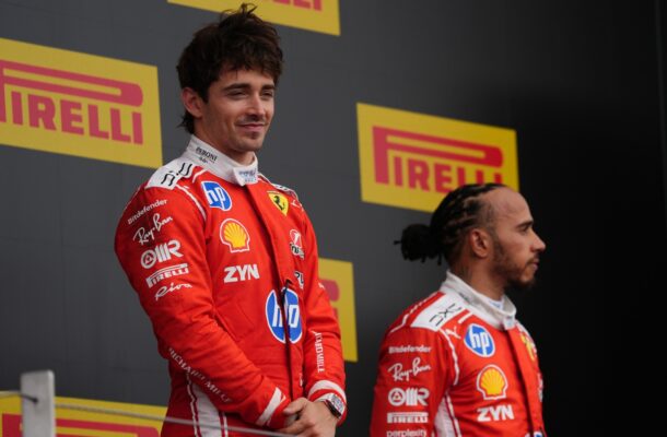 Two Ferrari drivers in red racing suits stand on a podium with yellow Pirelli signs behind them.