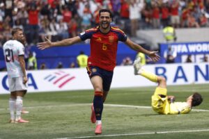 Spanish player in red jersey #6 celebrates with arms outstretched on the pitch as the goalkeeper lies on the ground behind him.