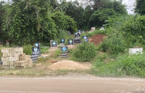 Row of small black tombstones with blue circular emblems along a dirt roadside, with piles of bricks and dirt and dense greenery nearby.