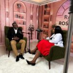 Two people sit in green armchairs on a pink-themed TV set, speaking into microphones during an interview.
