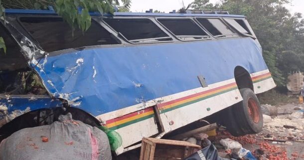 Blue bus overturned on its side with shattered windows and scattered debris; a few people are in the background.