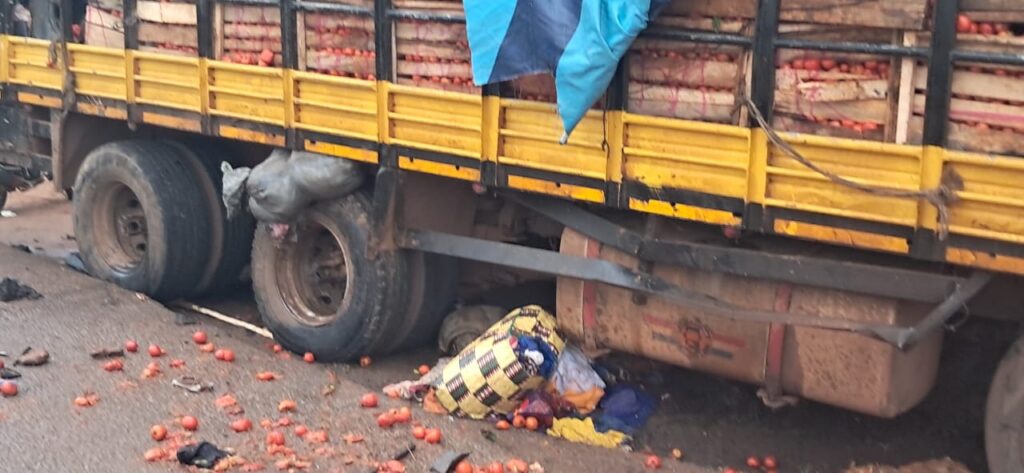 A large cargo truck with yellow side rails on a road; red tomatoes are spilled from crates under the trailer and scattered across the pavement.