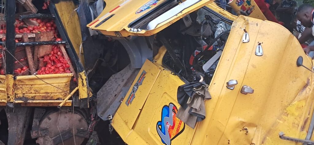 Two yellow trucks crash; a tomato trailer on the left is spilling red tomatoes into the scene, with the other cab crumpled and open nearby.