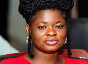 Close-up portrait of a woman with braided hair wearing a red top, looking at the camera with a neutral expression.