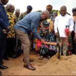 Officials and community members gather for a ground-breaking ceremony as a man in a blue shirt digs into a dirt mound with a shovel while others look on.