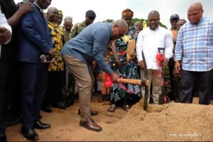Officials and community members gather for a ground-breaking ceremony as a man in a blue shirt digs into a dirt mound with a shovel while others look on.