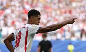Soccer player in a white jersey with red vertical stripes points to the right during a match, with a blurred stadium crowd behind him.