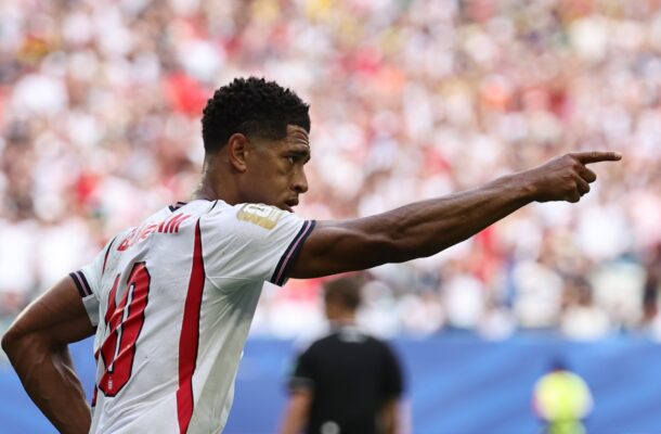 Soccer player in a white jersey with red vertical stripes points to the right during a match, with a blurred stadium crowd behind him.