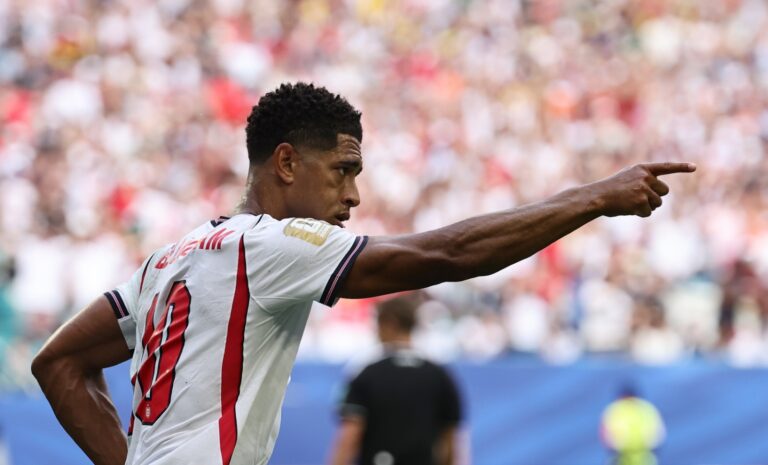 Soccer player in a white jersey with red vertical stripes points to the right during a match, with a blurred stadium crowd behind him.