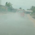 Red car emitting a thick cloud of smoke on a rural road, with pedestrians and palm trees along the roadside.