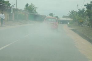 Red car emitting a thick cloud of smoke on a rural road, with pedestrians and palm trees along the roadside.