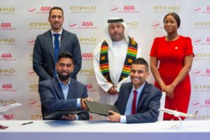 Two men in suits sign documents at a table during a ceremony, flanked by a man in traditional dress and a woman in red, with model airplanes and Etihad/AWA logos in the background