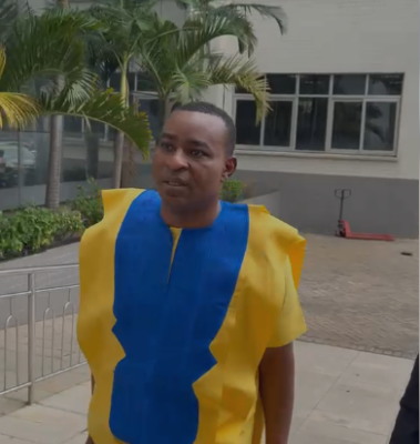 Wontumi court | Ghanaguardian.com Man standing outdoors wearing a bright yellow and blue traditional tunic, with palm trees and a building in the background.