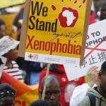 Crowd of protesters holding anti-xenophobia signs, including a yellow banner that says 'We Stand against Xenophobia' and other placards.