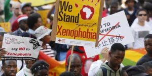 Crowd of protesters holding anti-xenophobia signs, including a yellow banner that says 'We Stand against Xenophobia' and other placards.