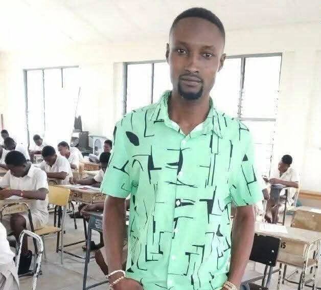 Portrait of a young man in a bright green geometric-pattern shirt standing in a classroom, with students seated at desks in the background.