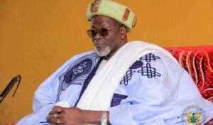 Elderly man in traditional West African clothing: white-blue agbada robe with blue patterns, green turban, sunglasses, seated with hands clasped.