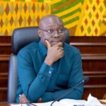 A man in a teal shirt sits at a desk in a formal meeting room, hand on chin, with a Ghana flag nearby and a colorful patterned backdrop.