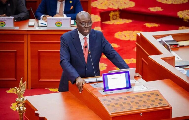 Man in a navy suit and red tie speaks at a wooden podium in a formal legislative chamber with a microphone nearby