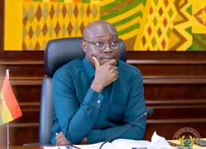 A man in a teal shirt sits at a desk in a formal meeting room, hand on chin, with a Ghana flag nearby and a colorful patterned backdrop.