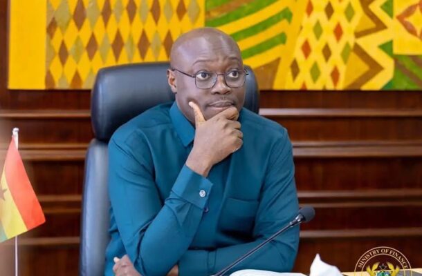 ato forson | Ghanaguardian.com A man in a teal shirt sits at a desk in a formal meeting room, hand on chin, with a Ghana flag nearby and a colorful patterned backdrop.