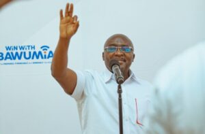 Man in a light shirt and glasses speaks into a microphone on a stage, his right hand raised in the air as he addresses an audience.