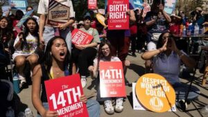 Diverse group of protesters at an outdoor rally holding signs about reproductive rights, including a front-left sign reading 'The 14th Amendment'.