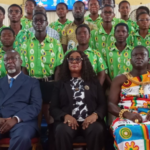 Group portrait at a formal event: five seated adults in front row, with a large crowd in matching green shirts behind them.