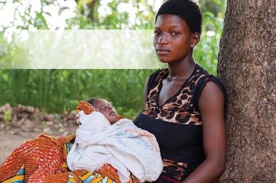 Mother sits under a tree holding her newborn swaddled in a white cloth with orange patterned fabric nearby, outdoors in a rural area.