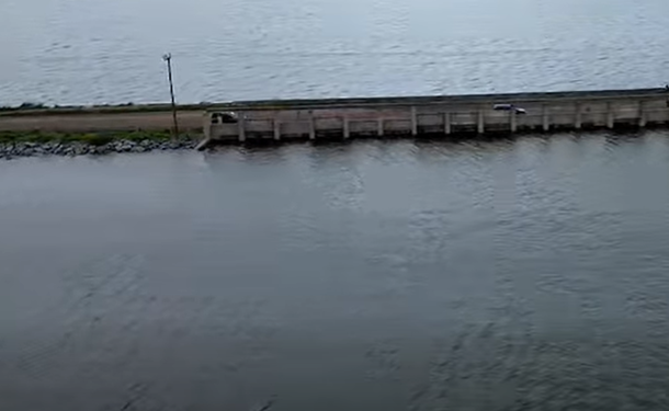 Long wooden pier or breakwater extends into calm water, with a single light post near its start and a grassy area beside.