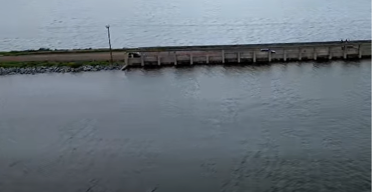 Long wooden pier or breakwater extends into calm water, with a single light post near its start and a grassy area beside.