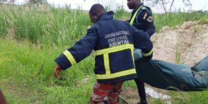 Two Ghana National Fire Service firefighters in dark jackets with yellow reflective bands carry a green casualty bag through tall grass in a field.