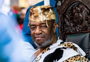 Man seated on an ornate carved chair wearing a fur crown and leopard-print cloak, looking at the camera.