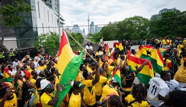 Large crowd wearing yellow shirts waving Ghana flags at an outdoor rooftop rally with a city skyline in the background.