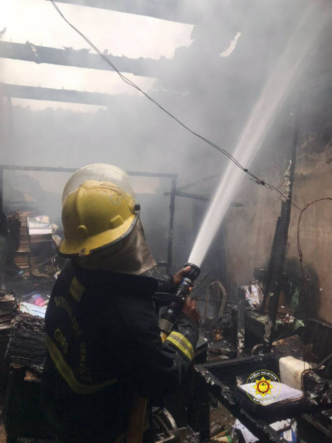 Firefighter in a yellow helmet sprays water to put out a smoke-filled, burned building interior.