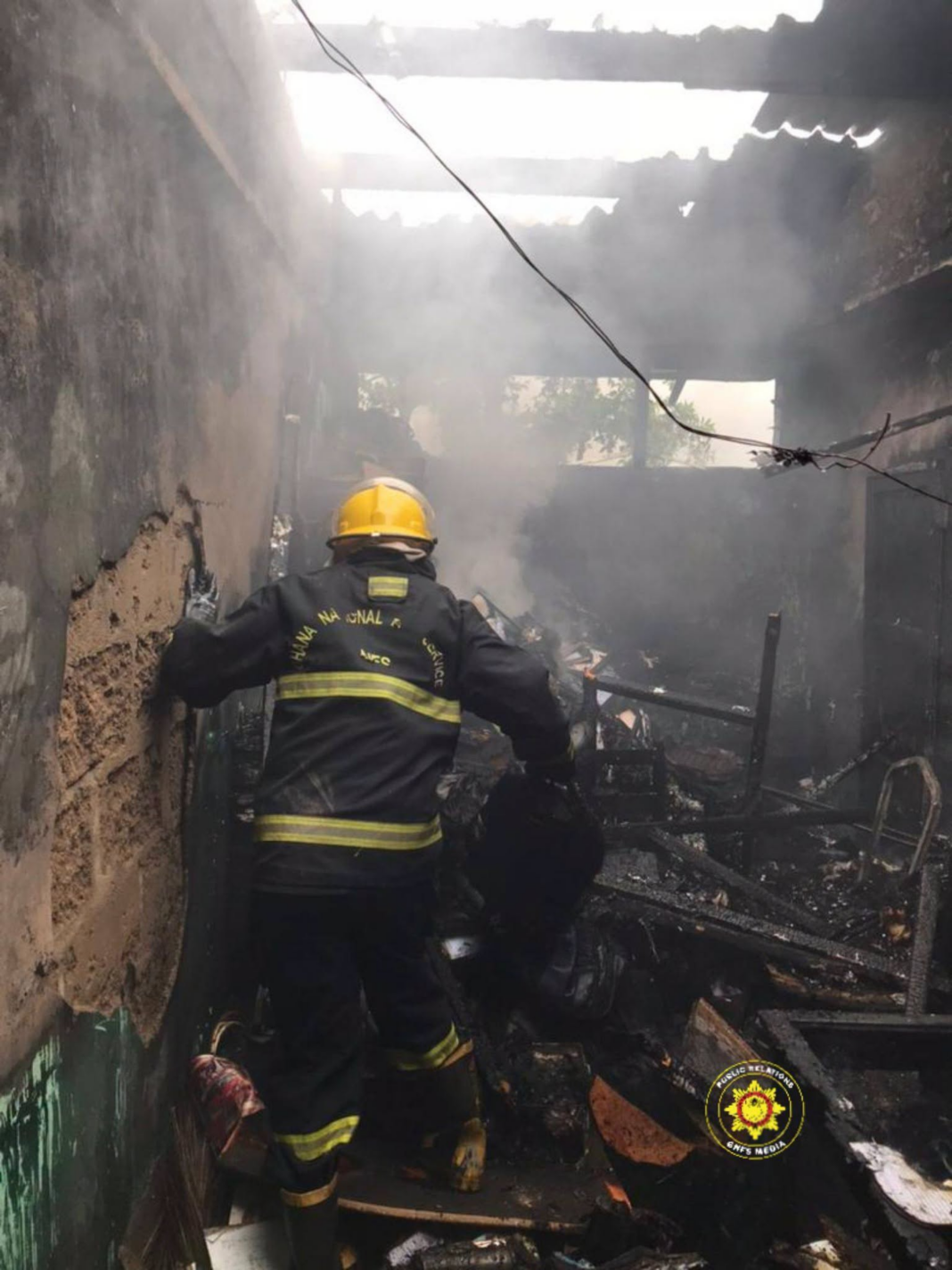 Firefighter in yellow helmet and reflective gear moves through smoky, charred rubble inside a burned building during a rescue operation.