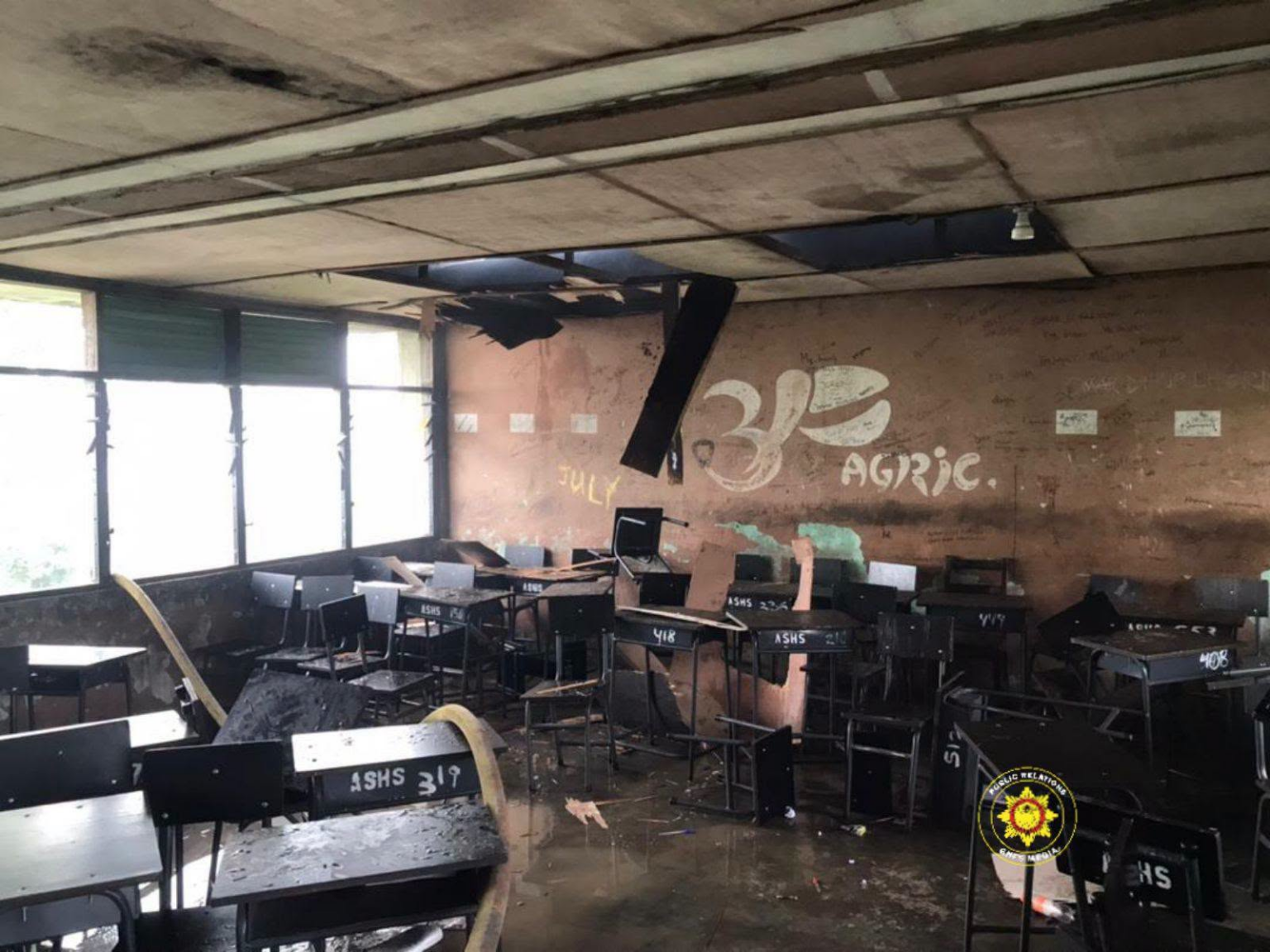 Abandoned, damaged classroom with broken ceiling panels, scattered desks, and graffiti on a brown wall.
